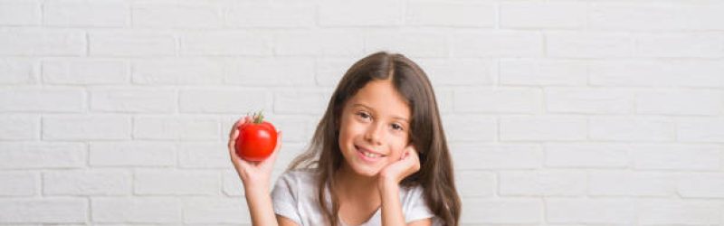 Young hispanic kid sitting on the table eating fresh tomato with a happy face standing and smiling with a confident smile showing teeth